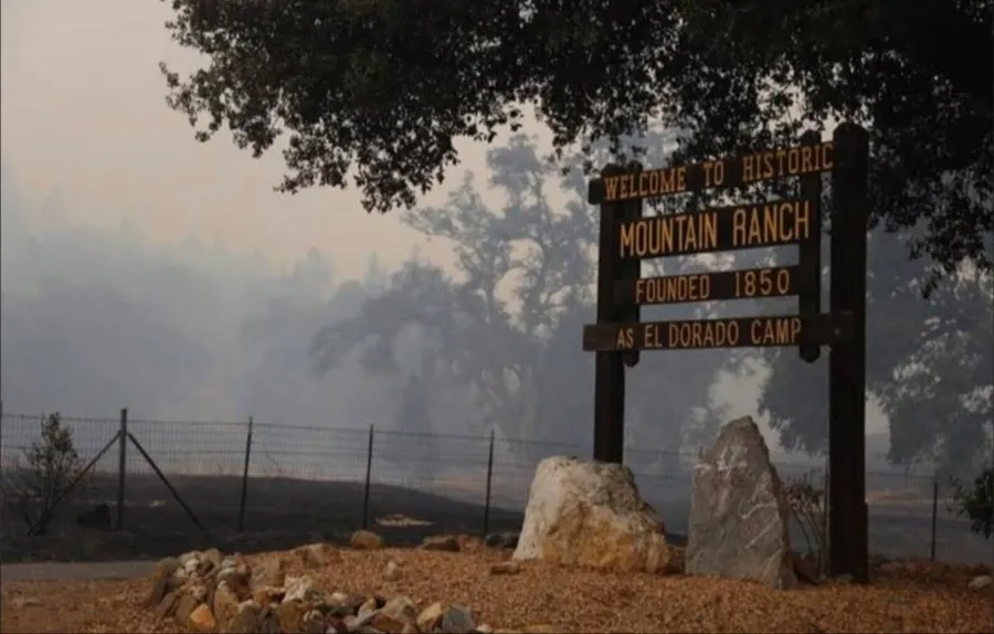 The Welcome to Historic Mountain Ranch sign standing in heavy smoke, with burnt ground behind the fence beside it and a green oak overhead